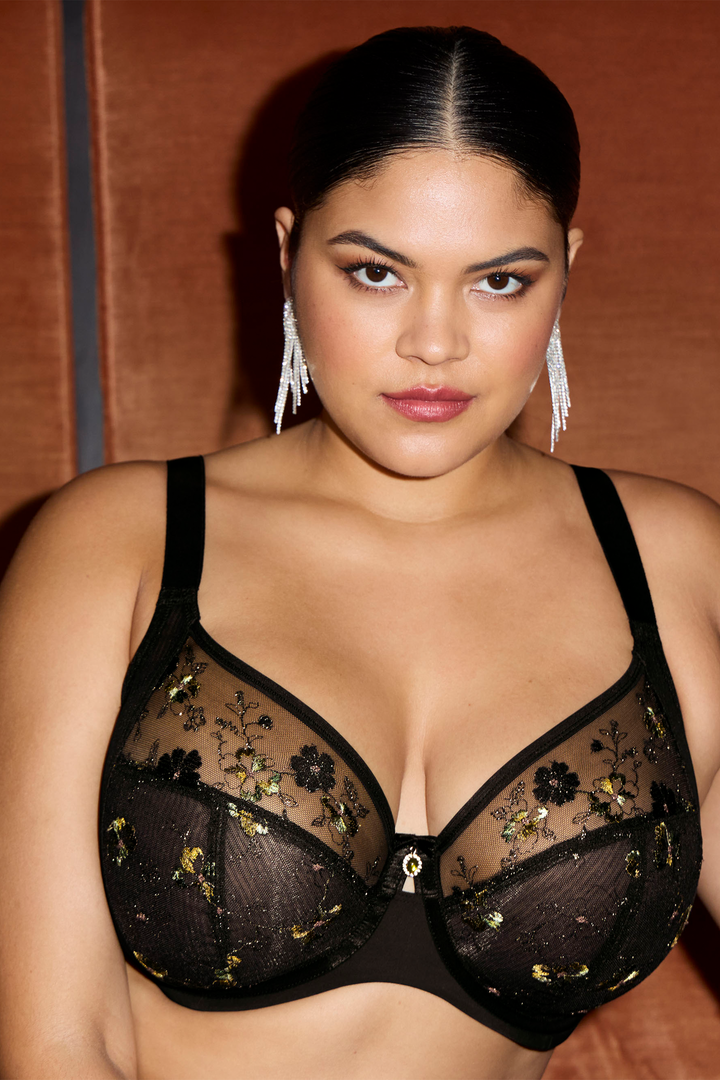 A woman wearing a black sheer floral lace bra with gold floral embroidery, photographed against a warm brown background.